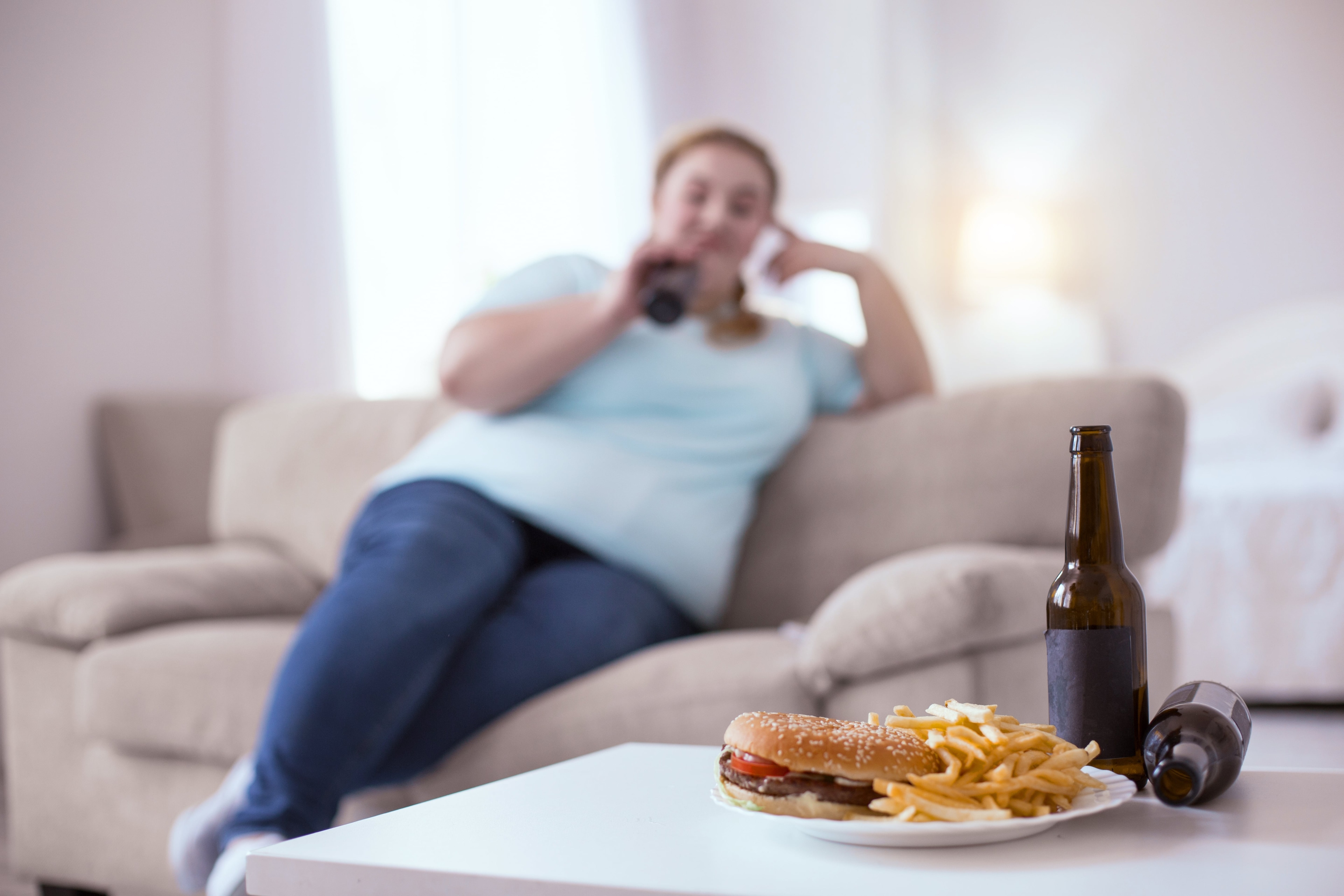 Túlsúlyos hölgy ül a kanapén, előtte sör és hamburger.